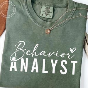 May include: A green t-shirt with the text "Behavior Analyst" in white lettering with a small heart symbol.