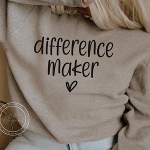 Peut inclure: Un sweat-shirt gris clair avec le texte "difference maker" en lettres noires et un petit symbole de cœur.