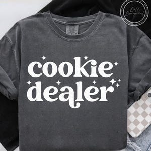 Op de afbeelding: Een donkergrijs sweatshirt met lange mouwen met de woorden "cookie dealer" in grote, witte, gestileerde letters. Witte sterren accentueren de tekst. De sweatshirt is gemaakt van een zacht materiaal.