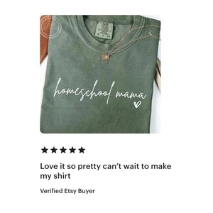 May include: An olive green t-shirt with the words "homeschool mama" in white script, a small heart, and a delicate necklace. The shirt is folded, and the text is centered. The shirt is a casual wear item.