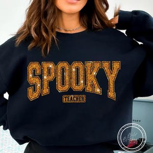 May include: Black sweatshirt with the word "SPOOKY" in gold glitter lettering. Below the word "SPOOKY" is the word "TEACHER" in a smaller gold glitter font. The sweatshirt is a crew neck style.