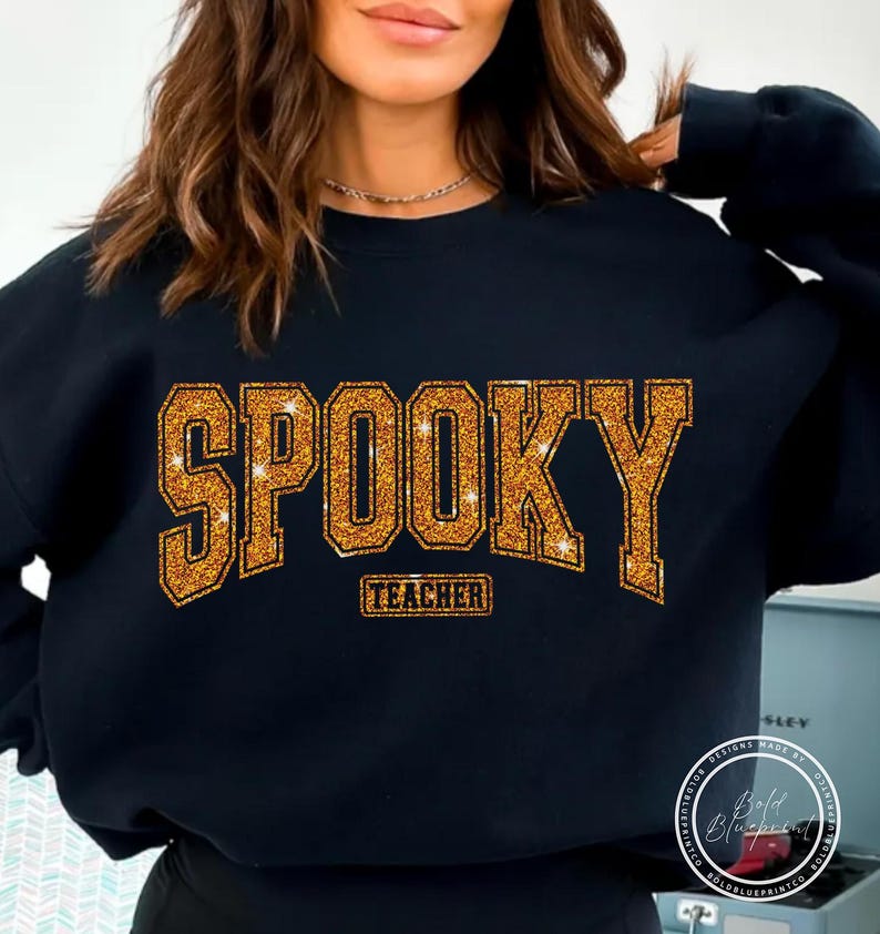 May include: Black sweatshirt with the word "SPOOKY" in gold glitter lettering. Below the word "SPOOKY" is the word "TEACHER" in a smaller gold glitter font. The sweatshirt is a crew neck style.