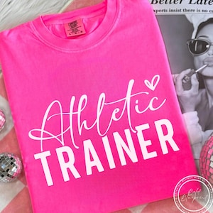 May include: A vibrant pink t-shirt featuring the text "Athletic Trainer" in white, cursive and block lettering. A small white heart is above the word "Trainer". The shirt is folded on a pink and white checked surface.