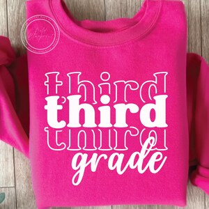 May include: A bright pink sweatshirt with the words "third grade" in white, stacked vertically. The text has a retro, layered design. The sweatshirt has a crew neck and long sleeves. A circular logo is visible in the upper left corner.