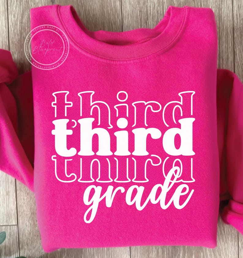 May include: A bright pink sweatshirt with the words "third grade" in white, stacked vertically. The text has a retro, layered design. The sweatshirt has a crew neck and long sleeves. A circular logo is visible in the upper left corner.