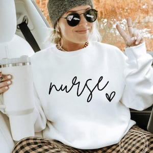 Puede incluir: Sudadera blanca con la palabra "nurse" escrita en cursiva negra con un pequeño corazón al lado.