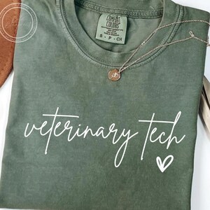 May include: An olive green t-shirt with the words "veterinary tech" in white script and a small heart. A gold-colored necklace with a pendant is draped over the shirt. The shirt has a Comfort Colors tag.