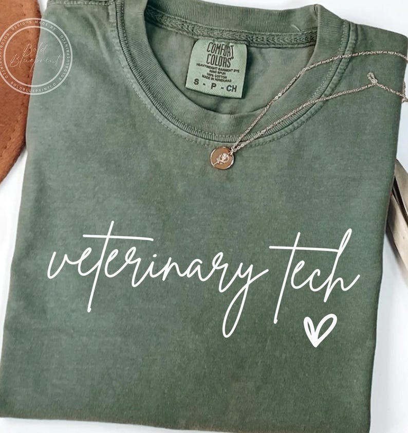 May include: An olive green t-shirt with the words "veterinary tech" in white script and a small heart. A gold-colored necklace with a pendant is draped over the shirt. The shirt has a Comfort Colors tag.