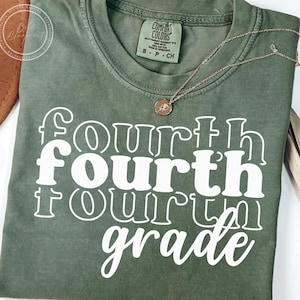May include: An olive green t-shirt with the words "Fourth Grade" in white, stacked vertically. The shirt has a crew neck and short sleeves. A gold-colored necklace with a small pendant rests on the shirt.