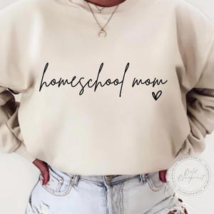 May include: Cream-colored sweatshirt with the words "homeschool mom" in black script, accompanied by a small heart. The sweatshirt is worn with light-wash denim jeans. A necklace with a pendant is visible.