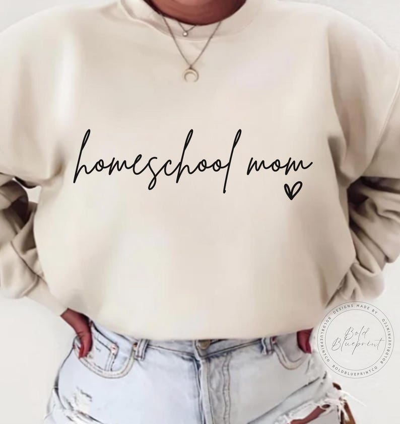 May include: Cream-colored sweatshirt with the words "homeschool mom" in black script, accompanied by a small heart. The sweatshirt is worn with light-wash denim jeans. A necklace with a pendant is visible.