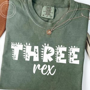 以下が含まれることがあります： 恐竜をテーマにしたフォントで「THREE rex」と書かれた白いテキストが施された緑色のTシャツ。Tシャツは使い古したような風合いがあり、柔らかく快適な生地で作られています。