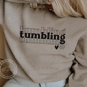 May include: A light gray sweatshirt with the word "tumbling" in a stacked, retro font, with a heart symbol. The sweatshirt is a neutral color and has long sleeves. The design is in black.