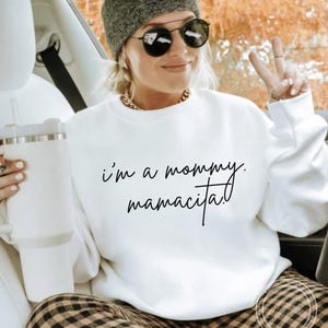 May include: White sweatshirt with the text "I'm a mommy. mamacita!" in black script. The sweatshirt is worn by a person wearing a gray beanie and sunglasses.