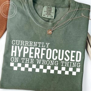 May include: An olive green t-shirt with the text "CURRENTLY HYPERFOCUSED ON THE WRONG THING" in white block letters. A silver necklace with a small pendant rests on the shirt. The shirt has a checkerboard pattern at the bottom.