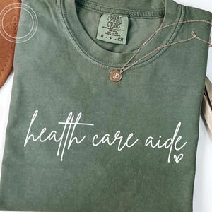 May include: An olive green t-shirt with the words "health care aide" in white script. A gold-coloured necklace with a small pendant rests on the shirt. The shirt is folded and has a Comfort Colors label.