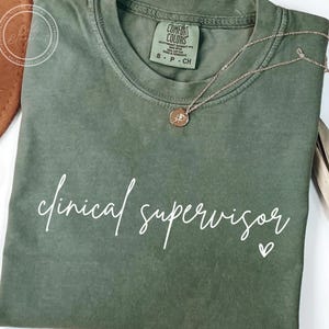 May include: Olive green t-shirt with the words "clinical supervisor" in white script. A small white heart is next to the text. A gold necklace with a pendant is on the shirt.