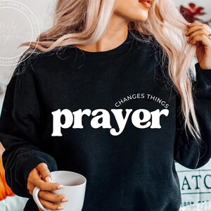 Peut inclure: Sweat-shirt noir avec le mot "prayer" en blanc, surmonté de "changes things". Une personne tient le sweat-shirt et une tasse blanche. Le sweat-shirt est de style col rond.