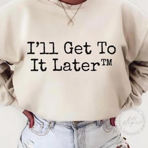 May include: A cream-coloured sweatshirt featuring the phrase "I'll Get To It Later" in bold, black lettering. The sweatshirt is worn with light-wash denim jeans. A necklace is also visible.