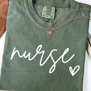 Peut inclure: Un t-shirt vert avec le mot "nurse" &eacute;crit en cursive blanche avec un petit c&oelig;ur &agrave; c&ocirc;t&eacute;. Le t-shirt porte une &eacute;tiquette indiquant "Comfort Colors".