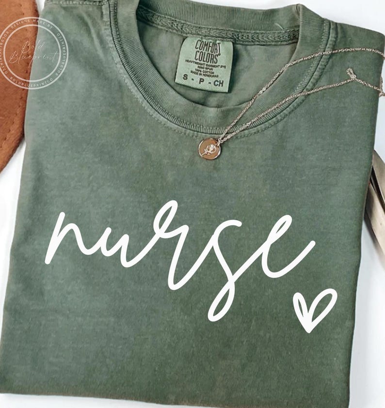 Peut inclure: Un t-shirt vert avec le mot "nurse" &eacute;crit en cursive blanche avec un petit c&oelig;ur &agrave; c&ocirc;t&eacute;. Le t-shirt porte une &eacute;tiquette indiquant "Comfort Colors".