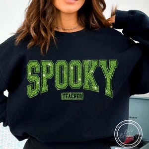 May include: Black sweatshirt with the word "SPOOKY" in large, glittery green letters. Below the word "SPOOKY" is the word "TEACHER" in a smaller, rectangular green font. The sweatshirt is a Halloween-themed item.