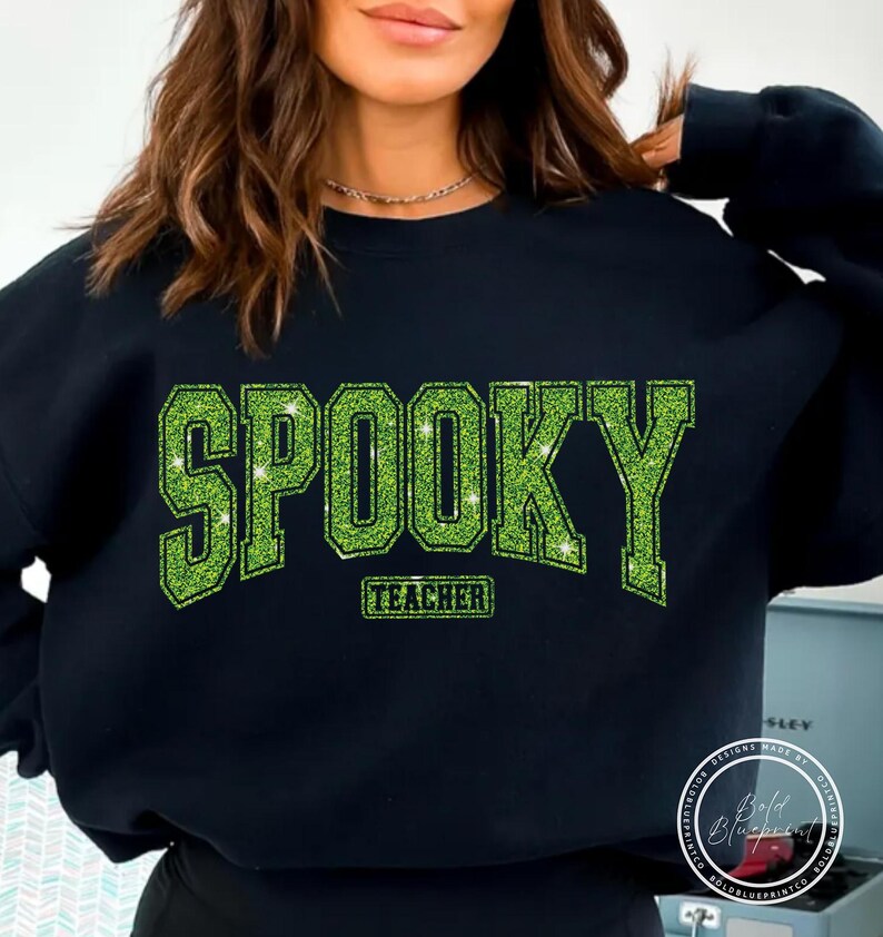 May include: Black sweatshirt with the word "SPOOKY" in large, glittery green letters. Below the word "SPOOKY" is the word "TEACHER" in a smaller, rectangular green font. The sweatshirt is a Halloween-themed item.