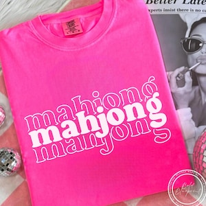May include: A bright pink t-shirt with the word "mahjong" stacked three times in white, with a retro font. The shirt is folded and lies on a pink and white checkered surface. The shirt has a crew neck and short sleeves.