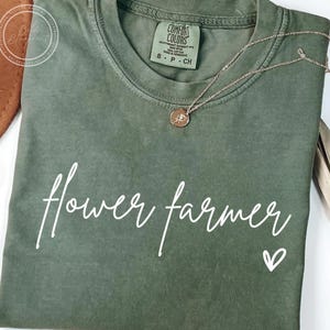 May include: A green t-shirt with the text "flower farmer" in white script and a small white heart. The shirt has a distressed look and is made of a soft, comfortable fabric.