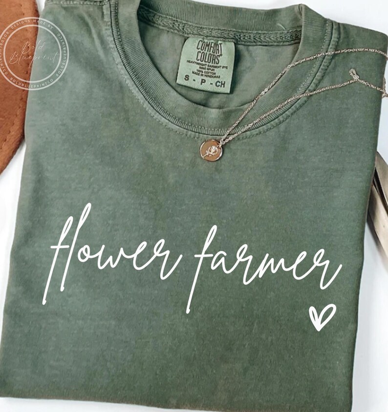 May include: A green t-shirt with the text "flower farmer" in white script and a small white heart. The shirt has a distressed look and is made of a soft, comfortable fabric.