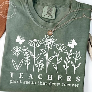 May include: A green t-shirt with a white graphic of flowers and butterflies. The text "TEACHERS plant seeds that grow forever" is printed on the shirt.