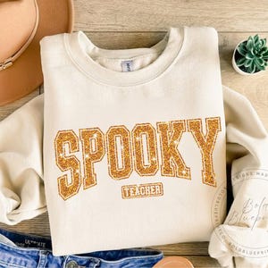 May include: Cream-colored sweatshirt with the word "SPOOKY" in gold glitter lettering. Below the word "SPOOKY" is the word "TEACHER" in a smaller gold glitter font. The sweatshirt is folded on a wooden surface with a hat and a small plant.
