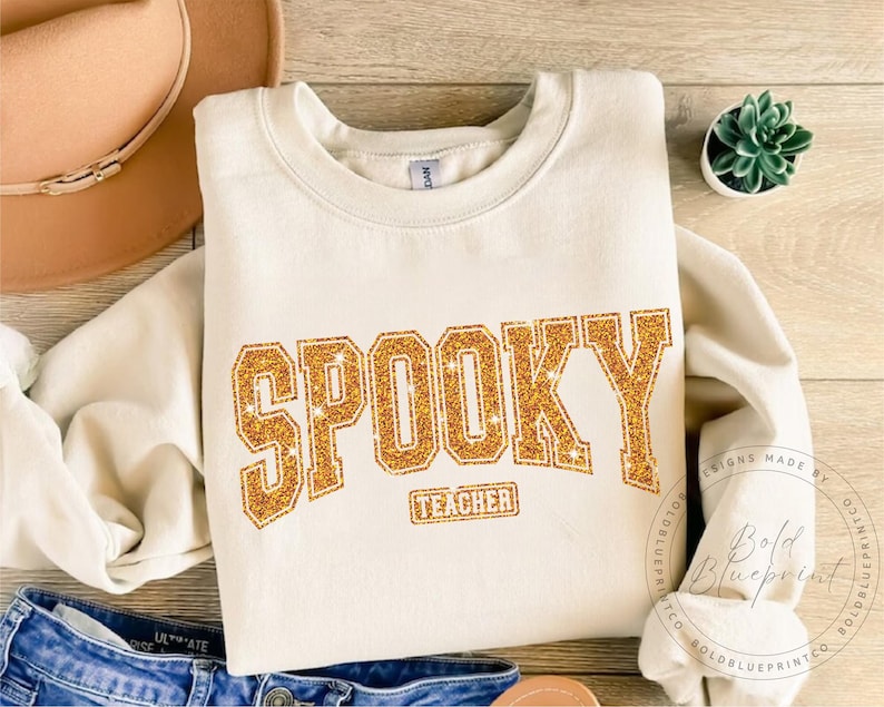 May include: Cream-colored sweatshirt with the word "SPOOKY" in gold glitter lettering. Below the word "SPOOKY" is the word "TEACHER" in a smaller gold glitter font. The sweatshirt is folded on a wooden surface with a hat and a small plant.