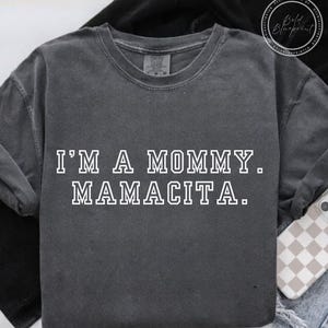 May include: Dark gray long-sleeve sweatshirt with the text "I'M A MOMMY. MAMACITA." in white block letters. The sweatshirt has a crew neck and a relaxed fit. A phone with a checkered case is visible.