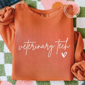May include: An orange-colored sweatshirt with the words "veterinary tech" in a cursive font and a small heart. The sweatshirt is made of a soft material and has a crew neck. The background is a colorful checkered pattern.