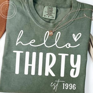 Puede incluir: Camiseta verde oliva con las palabras "hello THIRTY" en blanco, en escritura cursiva y letras de imprenta, con un pequeño corazón. El texto "est. 1996" está debajo. Un collar dorado con un colgante está sobre la camiseta.