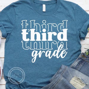 May include: A teal t-shirt with the words "third grade" in white, stacked vertically. The word "third" is in a bold, blocky font, while "grade" is in a cursive script. The shirt is made of a soft material and is likely for students.