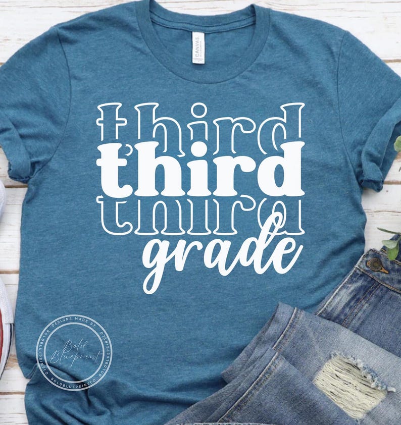 May include: A teal t-shirt with the words "third grade" in white, stacked vertically. The word "third" is in a bold, blocky font, while "grade" is in a cursive script. The shirt is made of a soft material and is likely for students.