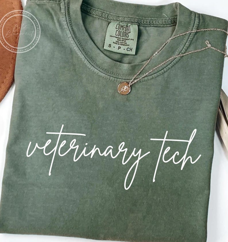 May include: Olive green t-shirt with the words "veterinary tech" in white script. A gold-colored necklace with a small pendant rests on the shirt. The shirt has a Comfort Colors tag.