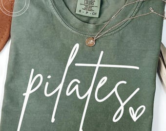 May include: Olive green t-shirt with the word "pilates" in white script and a small heart. A gold-colored necklace with a pendant rests on the shirt. The shirt has a Comfort Colors tag.