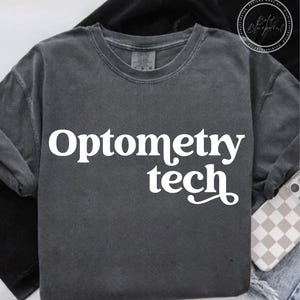 Puede incluir: Sudadera gris oscuro con cuello redondo con las palabras "Optometry tech" en blanco, con una fuente estilizada. La sudadera está hecha de un material suave y cómodo. Un teléfono con una funda a cuadros es visible en el fondo.