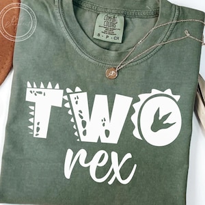 May include: Green t-shirt with white text that says "TWO rex" with a dinosaur footprint graphic. The shirt is made of a soft, comfortable material.
