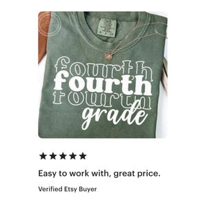 May include: Olive green t-shirt with the words "fourth grade" in a stacked, white, retro font. A gold necklace with a pendant rests on the shirt. The shirt is folded and displayed on a white surface.