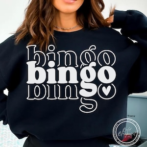May include: Black sweatshirt with the word "bingo" in large white letters. The word "bingo" is repeated twice, with a heart symbol at the end of the second word. The sweatshirt is a casual style.