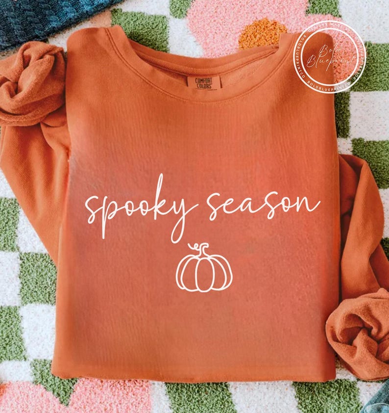 May include: An orange long-sleeved sweatshirt featuring the words "spooky season" in white script and a white pumpkin graphic. The sweatshirt is made of a soft material, ideal for autumn.