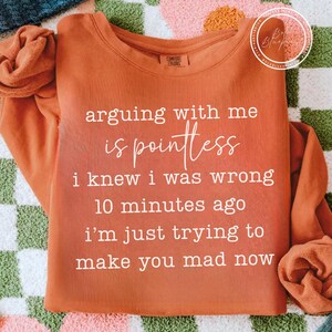 May include: Rust-colored long-sleeve sweatshirt with the text "arguing with me is pointless i knew i was wrong 10 minutes ago i'm just trying to make you mad now" in white script. The shirt has a crew neck and is made of a soft material.