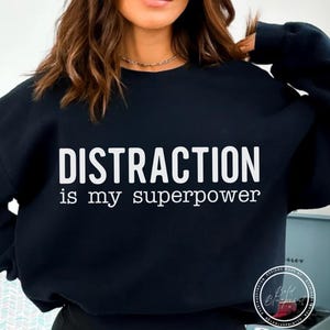 May include: Black sweatshirt featuring the white text "DISTRACTION is my superpower." The top has a crew neck and long sleeves. A circular logo is visible on the lower right side of the sweatshirt.