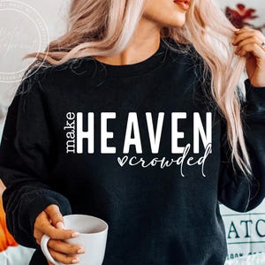 May include: Black sweatshirt with the text "make HEAVEN crowded" in white. The word "make" is stacked vertically on the left, with "HEAVEN" in large block letters and "crowded" in a script font below. A white mug is held in the hand.