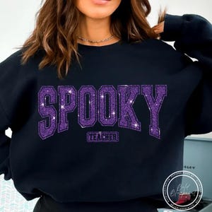 May include: Black sweatshirt with the word "SPOOKY" in large, glittery purple letters across the chest. Below the word "SPOOKY" is the word "TEACHER" in a smaller, rectangular font. The sweatshirt is a crew neck style.