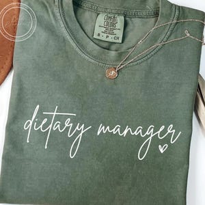 May include: An olive green t-shirt with the words "dietary manager" in white script. A gold-coloured necklace with a pendant rests on the shirt. The shirt is a Comfort Colors brand, size small.